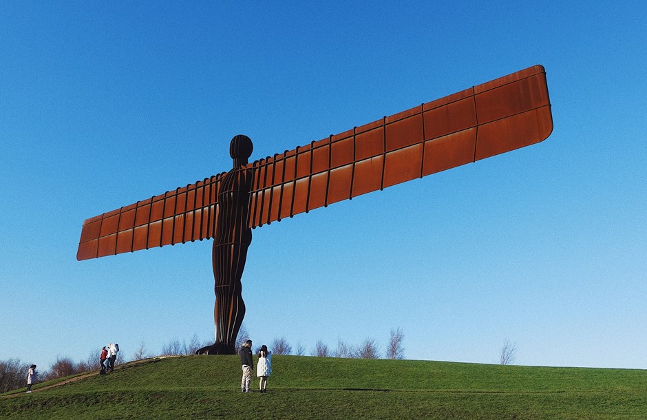 ‘Angel of the North’ artwork – monumental statue with long arms located on grass mound with some people milling about