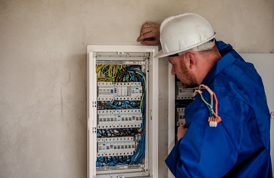 A worker in a hard hat and blue overall inspecting a fuse board